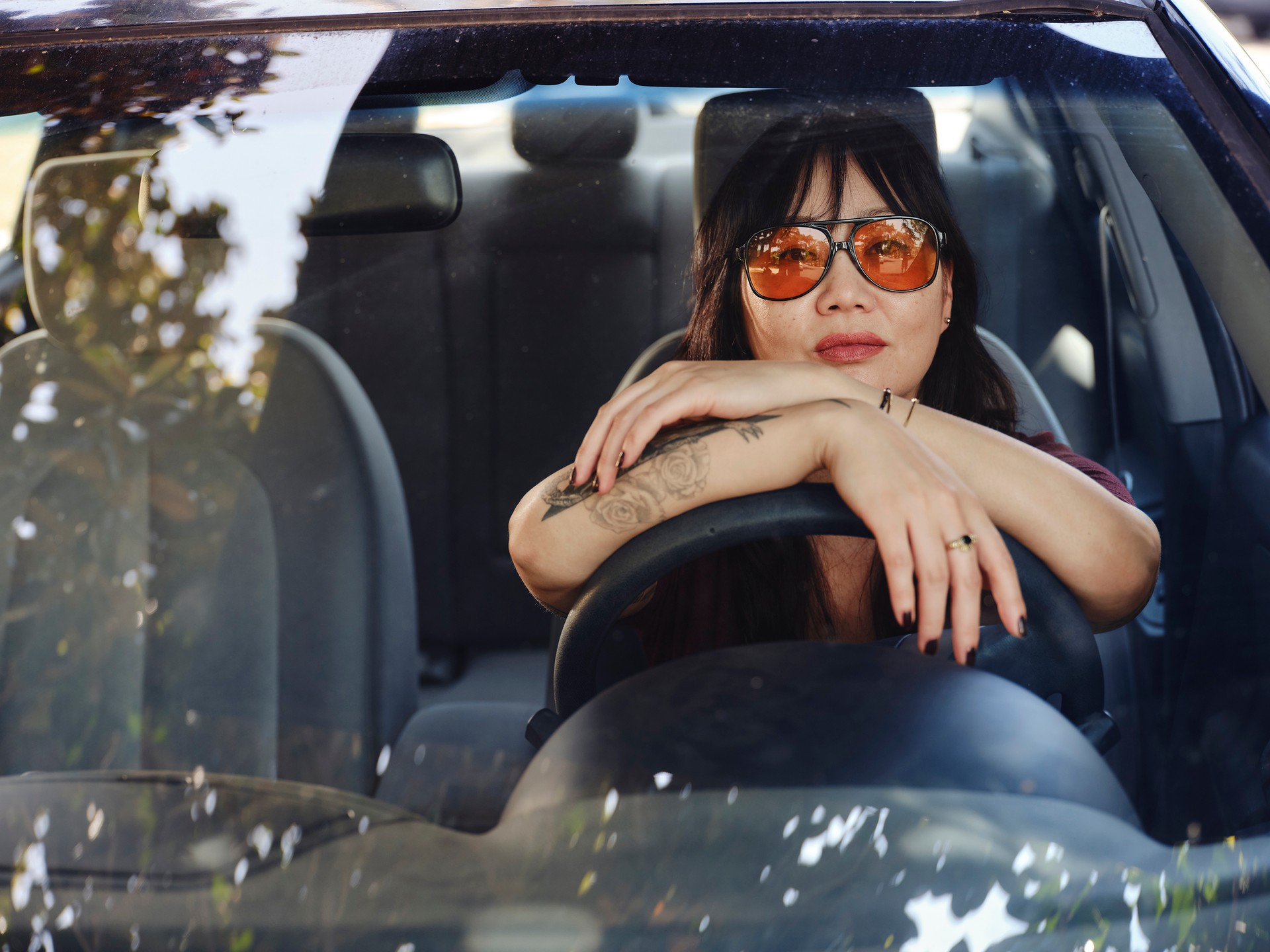 A hip Korean woman wearing orange-tinted sunglasses sits in her car with her arms draped over the steering wheel